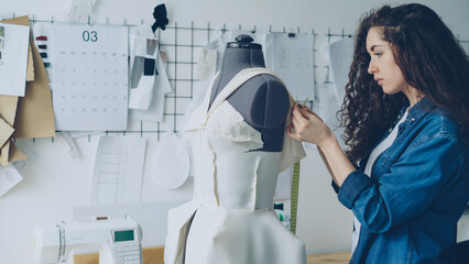 Young female tailor is adjusting clothes on tailoring dummy with sewing pins and measuring with...