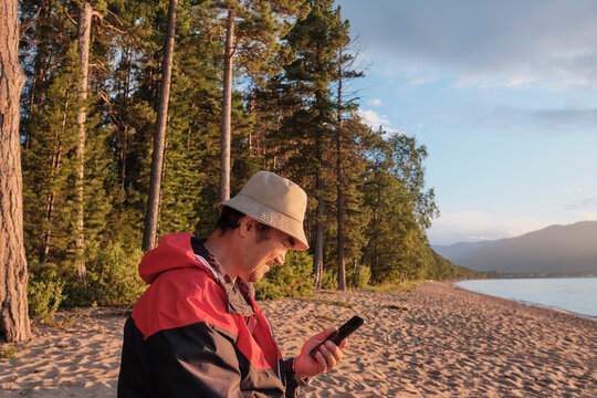 Senior asian man holding mobile phone on background of forest. Sandy beach of Lake Baikal, Russia