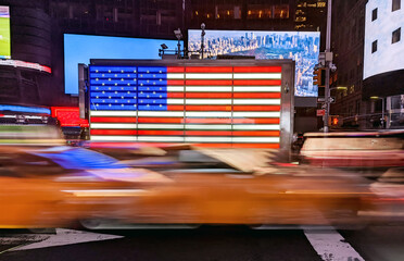 Yellow taxis driving past an American flag in Times Square, New York City with motion blur