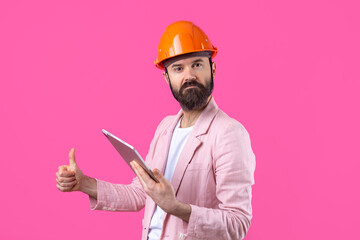 Portrait of young construction engineer wear orange hard hat, in a pink jacket standing on red studio background. A man with a tablet in his hands.