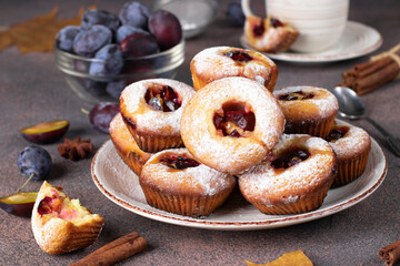 Homemade muffins with plums sprinkled powdered sugar on round plate on brown background