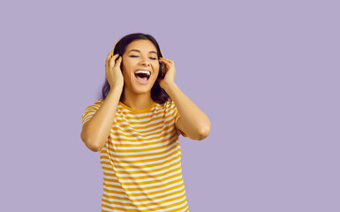 Positive woman in headphones listens to music and sings loudly isolated on pastel purple background. Smiling young woman in yellow striped T-shirt and wireless headphones sings with her eyes closed.