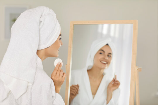 Beautiful woman in white towel turban on head holding cotton disk, looking at reflection in mirror and putting dermatological toner on face to prepare skin for moisturizing. Beauty, skin care concept