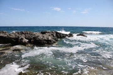 Waves crash over a rock in an aqua blue ocean with a blue sky on top.  Image has copy space.
