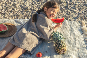 Little girl eats fruit lying on a blanket on the beach.