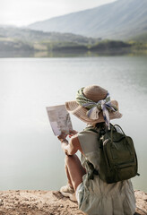 rear view of a woman in a hat sitting on the shore of a lake with a map in her hand. concept of travel and adventure.