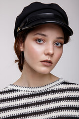 High fashion photo of a beautiful elegant young woman in a pretty black long leather skirt, black and white striped sweater with sleeves, hat posing over white, soft gray background. Studio Shot. 