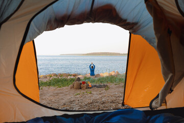 view from a tent on the shore of a lake or sea, a woman collects firewood or does yoga and...