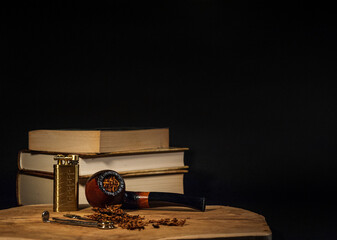 A tobacco pipe lying on a piece of wood with a pipe tamper, lighter and scattered tobacco with books in the background - obrazy, fototapety, plakaty