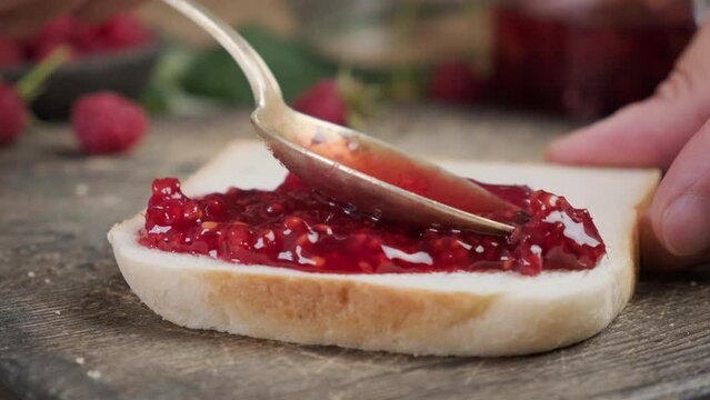 Spoon spreading raspberry jam on piece of toast. Preparation of sweet yummy sandwich for tasty breakfast. close up 4k video
