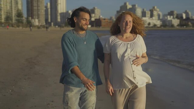 Happy couple. Pregnant redhead woman and boyfriend walking by sandy beach front to the ocean in Punta del Este Beach in Uruguay