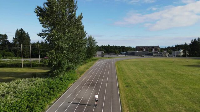 Young man skating along running track around a football field in a scenic location with the pacific ocean in background. Aerial pull away revealing a picturesque view of Canada's sunshine coast