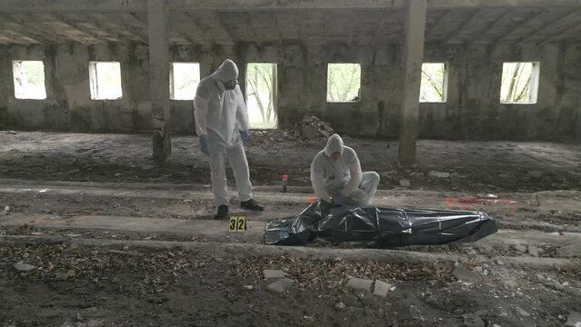 Two forensic specialists gather evidence and put the corpse in a black body bag. Dolly shot