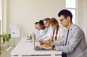 Serious busy people working in modern office workplace. Group of diverse employees sitting in row at white desk, working on laptop computers, reading business reports, taking notes. Side profile view