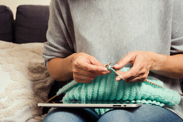 Middle age woman at home look at digital tablet computer screen and knitting sweater. Woman...