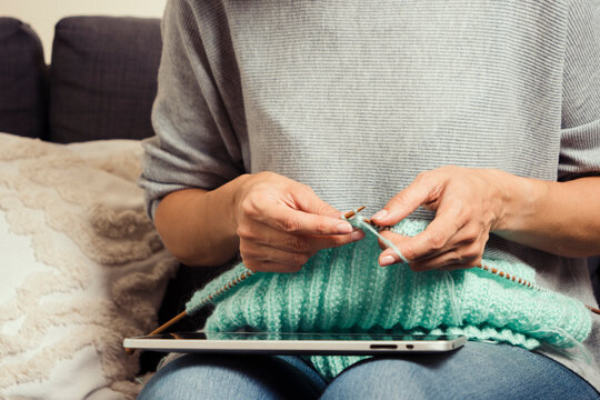 Middle Age Woman At Home Look At Digital Tablet Computer Screen And Knitting Sweater. Woman Learning Knitting, Watching Video Tutorial, Online Class Lesson, Follow Knitting Patterns