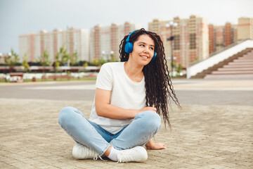 A smiling girl with dreadlocks in headphones is sitting on the floor in the park. A happy young woman relaxing with headphones, enjoying music. Space for copying.generation z