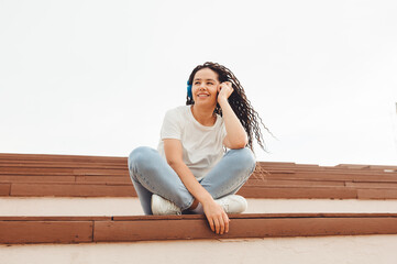 Portrait of a smiling young woman with dreadlocks sitting on a park bench and listening to music. Listening to music with headphones .generation z.