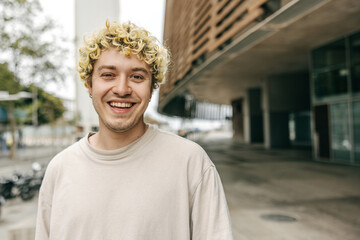Happy caucasian young guy broadly smiling for camera staying near university library. Curly blonde wear beige t-shirt. Concept human emotion.