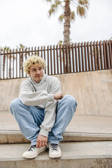 In full growth young caucasian guy with tattoo on face posing for camera sitting outdoors. Blonde hair man wears grey sweater and casual jeans. Concept stylish vacation, rest time.