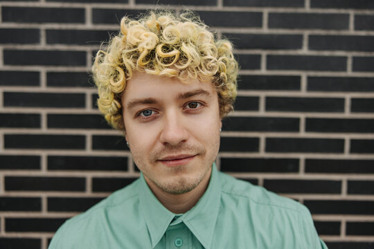 Portrait of young caucasian man with different eyes and tattoo on his face, standing black brick wall in background. Curly blond man wearing casual clothes looks at camera. Conceptual portrait.