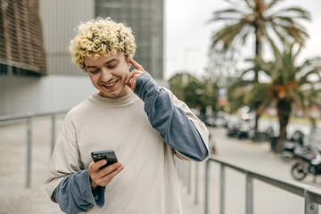 Portrait of handsome caucasian young guy wearing casual clothes near city mall. Curly blonde using smartphone on urban street. Concept technologies.