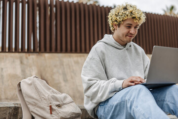 Stylish young caucasian guy with tattoo on face using laptop at street. Handsome man with backpack...