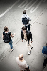 Young European man, wearing black jacket, light color pants, walking on sidewalk on street in New York City, passing by people, looking around.