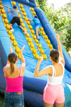 Young And Adult Men Using Sticks To Climb Up On Slide In Adventure Park. Women Supporting Them, Raising Hands And Gesturing.