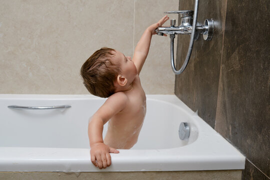 Happy toddler baby is playing in white bathtub with water faucet. Kid aged one year and two months