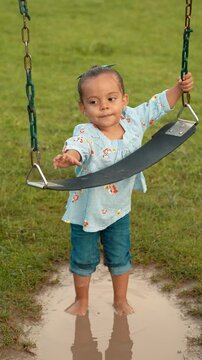 Ni&ntilde;a morena latina peque&ntilde;a moj&aacute;ndose en charco de lluvia jugando en columpio de parque en la naturaleza en un hermoso d&iacute;a al atardecer en exterior