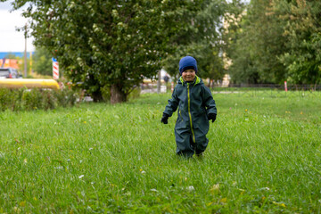 
A two-year-old boy runs through a forest park in the city on an autumn street in overalls and a hat

