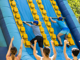 Two male friends having fun in outdoor amusement park on summer day, climbing on inflatable slide...