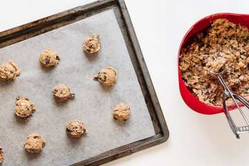 Round raw oatmeal raisin cookie dough scoops on sheet with baking paper made of flour, rolled oats, sugar, eggs, raisins, dried fruit, baking soda before baking next to bowl with dough and scoop - 531552635
