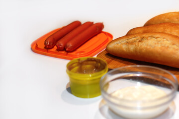 Defocus ingredients for making homemade hot dogs. Sausages in orange plate, fresh baked buns, mustard and sauce, served on white background.  Hotdog with different ingredients. Out of focus
