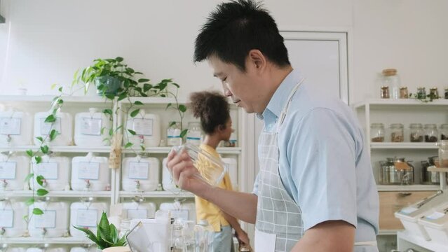 Asian male shopkeeper works with laptop, checks purchase order, a customer chooses natural organic product at display shelves in refill store, eco-friendly and plastic free shop, reusable container.