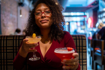 African American woman with curly black afro hair  wearing a red dress is drinking two mixed drinks at a bar.