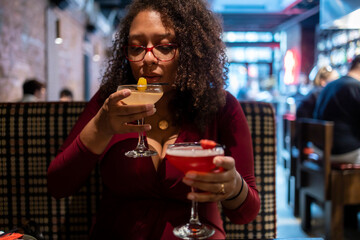 African American woman with curly black afro hair  wearing a red dress is drinking two mixed drinks at a bar.