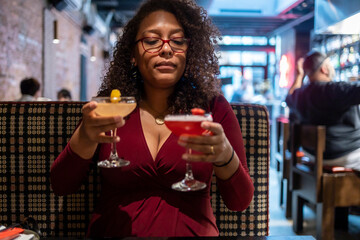 African American woman with curly black afro hair  wearing a red dress is drinking two mixed drinks at a bar.