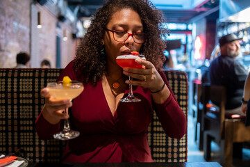 African American woman with curly black afro hair  wearing a red dress is drinking two mixed drinks at a bar.