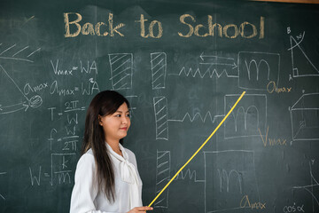 Back to school concept. Asian female teacher smiling with wooden stick pointing to blackboard at school in classroom, Happy beautiful young woman standing hold pointer to back board