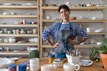 Happy joyful young Turkish woman successful self-employed potter wearing apron behind table with pottery tools smiling at camera, cheerful female ceramist posing against display of handmade ceramics - obrazy, fototapety, plakaty