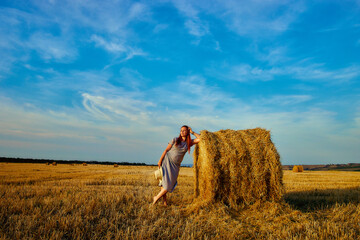 A beautiful young woman in a hat and a summer dress is sitting on a sheaf of hay in a field. Rural nature, wheat field, rest in the country, unity with nature