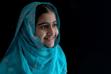 Naklejka premium Little Muslim girl with beautiful eyes wearing blue hijab smiling with someone, black background, side view