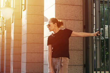 Profile image of a young woman in sunglasses holding on a fence rail behind her back on a sunlit street.