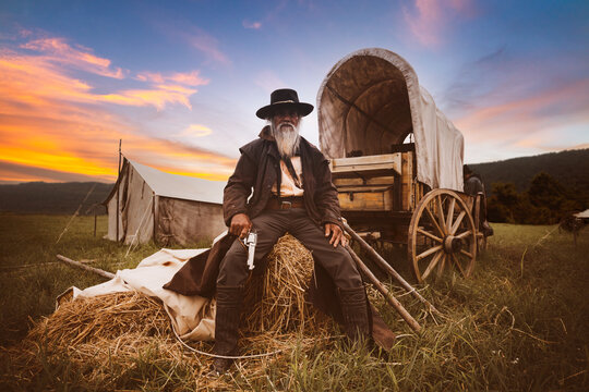 Oldest smart cowboy man wearing western style suite with cowboy hat holding gun on hand sit on haystack with horse carrier and tent is vintage 1800s life style concept.