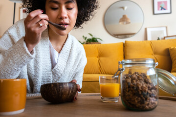 African American woman eating granola for breakfast at home living room. Coffee and orange juice,...