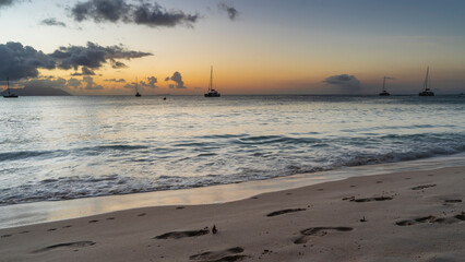 Romantic sunset on a tropical island. Blue clouds in the sky, highlighted in orange. Silhouettes of yachts on the horizon. Foam waves on the beach. Footprints in the sand. Seychelles. Mahe.