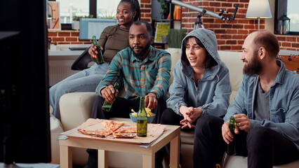 Diverse people watching film or tv show at home gathering, having fun with comedy or action movie. Using television channel program to enjoy cinema series with beer bottles at hangout.