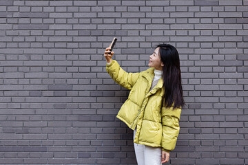 Fototapeta na wymiar Stylish Young Asian beautiful woman using smart phone for in outdoor cafe near gray brick wall at spring or autumn season. Blogger streaming broadcast for social media and communicating with followers